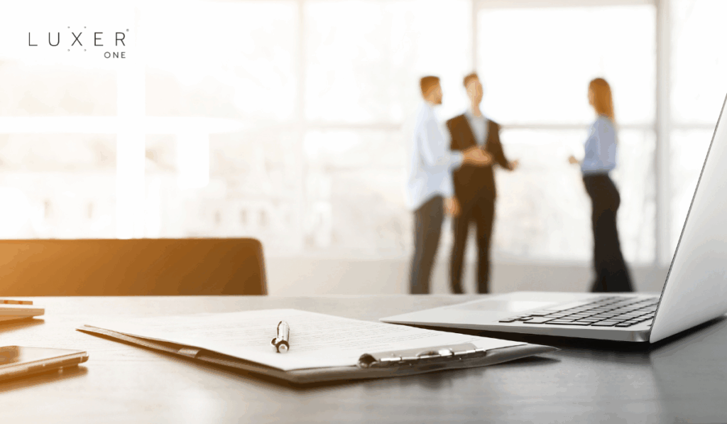 Laptop and clipboard on a desk with colleagues talking in the background in an office